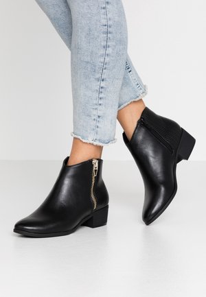 Sorte læderankelstøvler med sidelommer med lynlås, båret med lyseblå cropped jeans på en hvid gulv og ensfarvet baggrund.