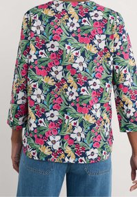 Bageste udsigt af person iført langærmet skjorte med et levende blomstermønster i hvid, pink, gul og grøn, kombineret med blå denim jeans.