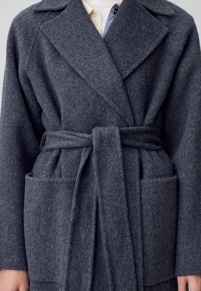 Abrigo de lana gris oscuro con diseño envolvente, amplias solapas, un cinturón y bolsillos frontales. Tiene una textura suave y una forma estructurada.