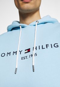 Světlomodrá mikina s kapucí s předním logem „TOMMY HILFIGER“ v tmavě námořnické modři a obdélníkovým červeno-bílo-černým znakem. Bílé šňůrky na stažení.