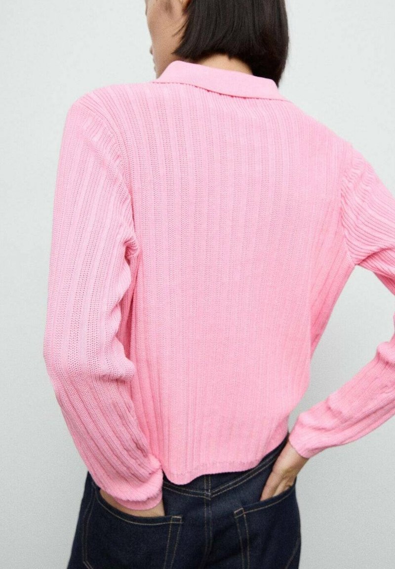 Personne aux cheveux courts et foncés portant un haut rose à manches longues côtelé et un jean foncé, les mains dans les poches arrière, face à un mur gris clair.