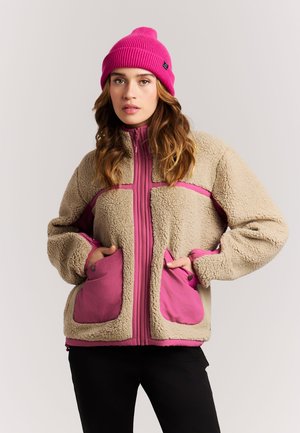 Woman wearing a beige and pink fleece jacket with front pockets and a bright pink knit beanie, standing with hands in pockets.