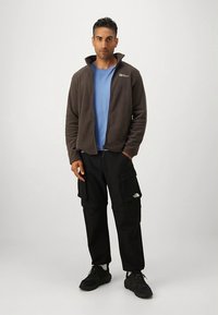 Brun fleecejakke, blå t-shirt, sorte cargobukser med lommer, mørke sneakers. Afslappet udendørs påklædning med funktionelle designdetaljer.