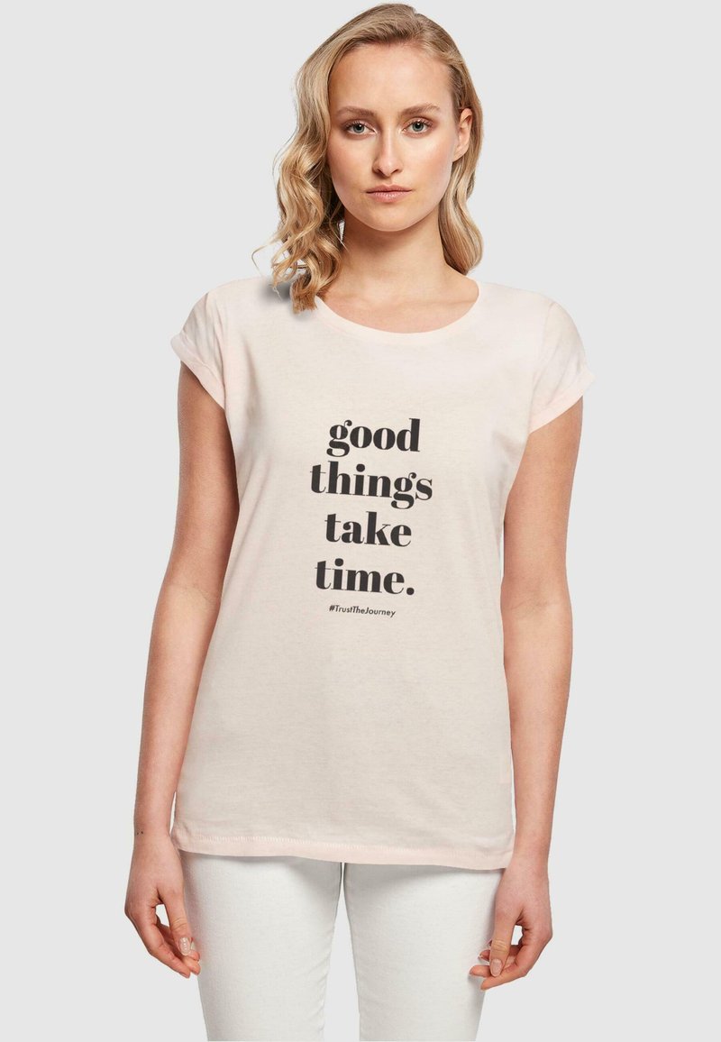 Lyserød t-shirt med korte ærmer lavet af bomuld, med sort tekst: "gode ting tager tid. #TrustTheJourney" fremtrædende vist.
