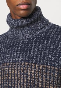 Pull en maille à col roulé bleu foncé avec des accents gris clair et beige, présentant un motif texturé et un design de col haut.