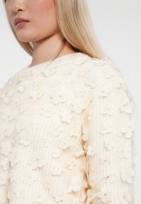Maglione color crema caratterizzato da un tessuto texturizzato con applicazioni floreali sparse e dettagli di leggero scintillio. Collo rotondo e maniche lunghe.