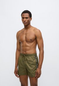 Oliivirohelised ujumispüksid elastse vöökohaga, nööriga ning musta värvi külglogo detailidega. Sile ja kerge kangas.