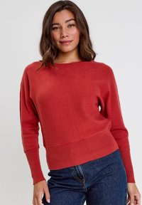 Suéter de punto color óxido con un escote amplio, puños y dobladillo acanalados. Corte ajustado, textura suave. Modelo combinado con jeans oscuros de cintura alta.