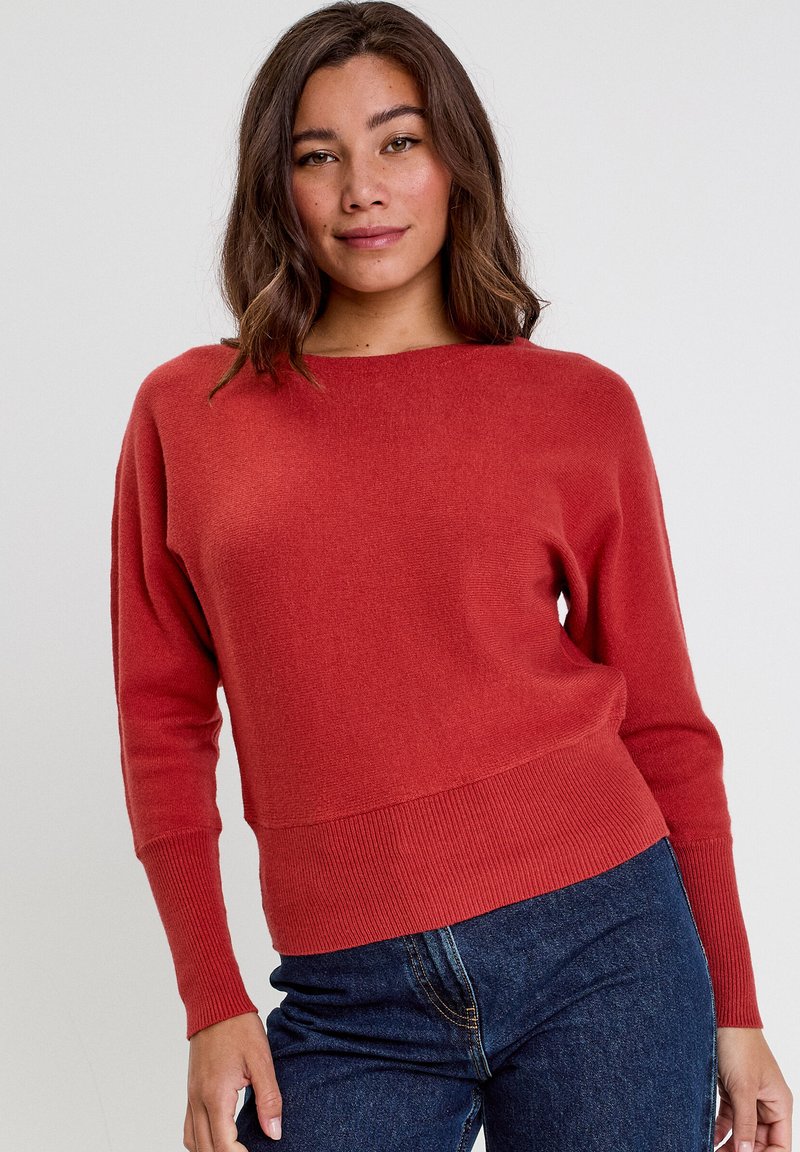 Rust red, knitted sweater with a wide neckline, ribbed cuffs, and hem. Tapered fit, soft texture. Model paired with high-waisted dark jeans.
