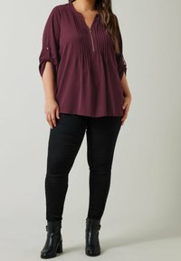 Femme portant un blouse prune à manches longues avec des plis et une fermeture éclair, un pantalon skinny noir et des bottines noires à talons blocs.