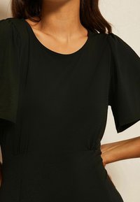 Mustapuku, jossa on lyhyet puffihihat, pyöreä pääntie ja sileä, teksturoitu kangas, jossa on hienovaraisia poimintoja vyötäröllä.