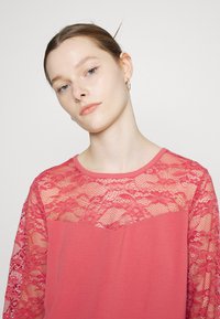 Top rosa com sobreposição de renda nas mangas e na parte superior do corpete. Mangas longas, decote redondo e textura suave.