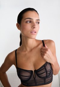 Czarny przezroczysty biustonosz o usztywnionej konstrukcji z miękkimi miseczkami, regulowanymi ramiączkami i detalem w kształcie dziurki z przodu. Tekstura jest delikatna i lekka.