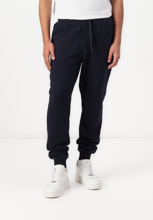Pantalones de chándal de algodón azul marino con cintura elástica, cordones y puños elásticos, combinados con zapatillas blancas.