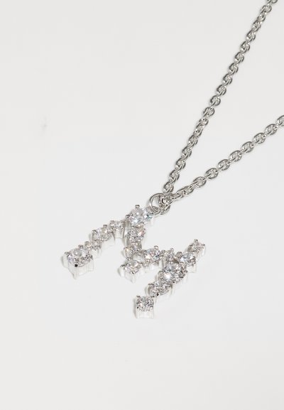 Ασημένια αλυσίδα κολιέ με μενταγιόν σε σχήμα 'M', πλαισιωμένο από διαυγείς πολύτιμους λίθους σε σχεδίαση με προεξοχές, προσφέροντας λάμψη και αντίθεση με το μέταλλο.