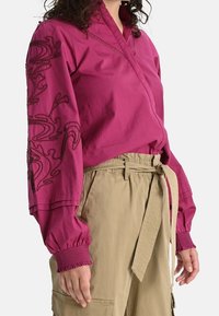 Blusa avvolgente fucsia con maniche lunghe e motivi ricamati a spirale, abbinata a pantaloni cargo beige chiaro con cintura. Tessuto dalla consistenza liscia.