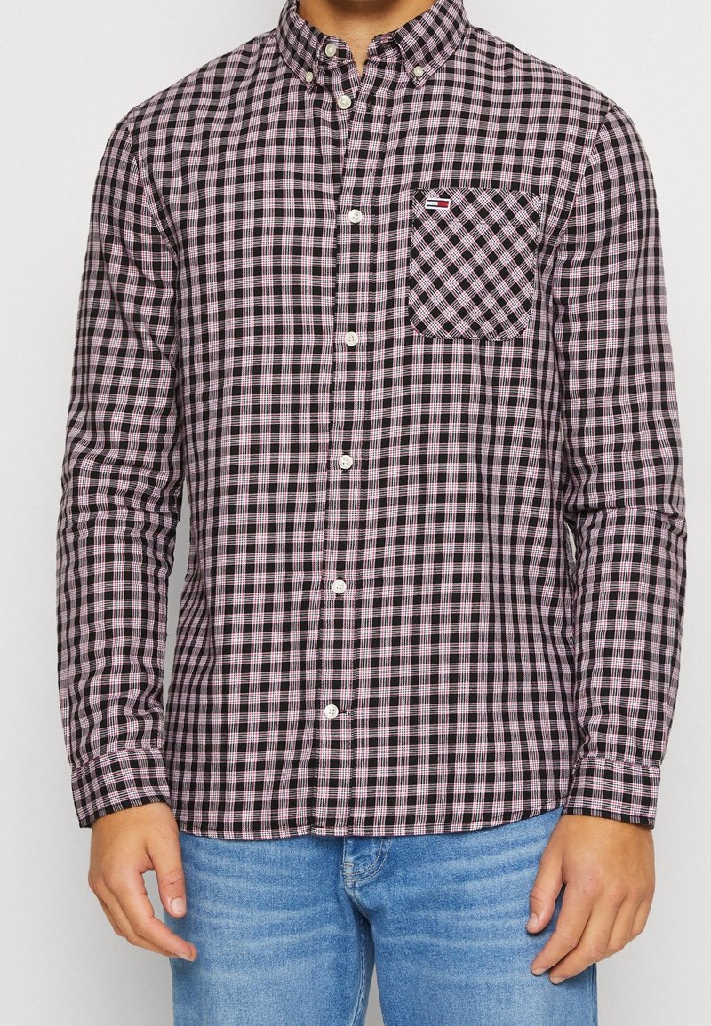 Homme portant une chemise à manches longues à carreaux noirs et roses avec poche poitrine et un jean bleu, debout devant un fond uni.