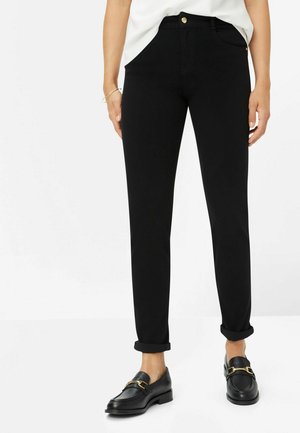 Pantaloni neri elasticizzati con una vestibilità slim, caratterizzati da vita alta, tasche frontali e orli arrotolati, abbinati a mocassini neri con dettagli dorati.