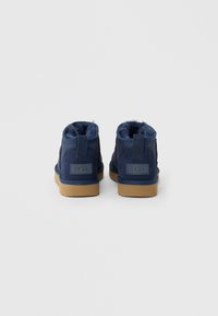 Μποτάκια UGG σε ναυτικό μπλε χρώμα με επένδυση από shearling και καφέ λαστιχένιες σόλες, φωτογραφημένα από πίσω σε καθαρό λευκό φόντο.