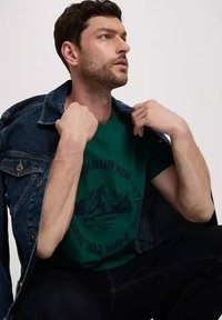 Zielona koszulka graficzna z motywem górskim i napisem, warstwowa pod ciemną dżinsową kurtką. Model pokazujący profil boczny.