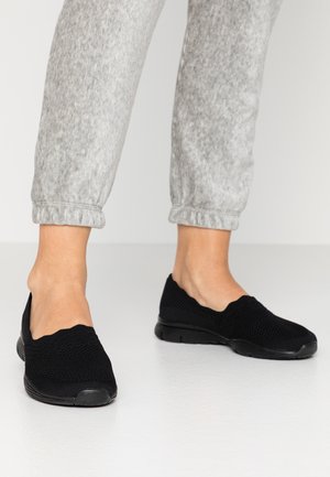 Pies con zapatillas negras de malla sin cordones combinadas con pantalones deportivos gris claro con puños, sobre un suelo blanco liso.