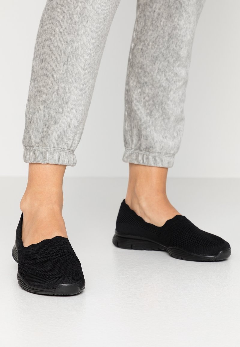 Pieds portant des baskets noires à enfiler en maille, associées à un pantalon de survêtement gris clair retroussé, sur un sol blanc lisse.