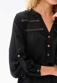 Schwarze Bluse mit V-Ausschnitt, Spitze-Akzenten und Knopfdetails. Lange Ärmel mit geknöpften Manschetten und einer Brusttasche.