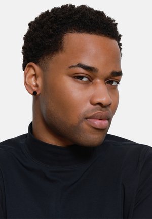 Zwarte hoge nek top van gladde stof. Korte, krullende haren. Zwarte studs als oorbellen. Neutrale achtergrond, focus op gezichtskenmerken en shirt.