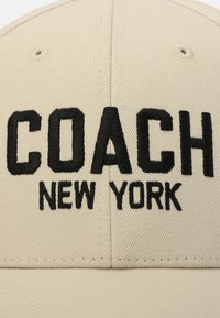 Beige keps med svart broderad text "COACH NEW YORK." Har en strukturerad design med ett texturerat tyg och ett ventilationshål på toppen.
