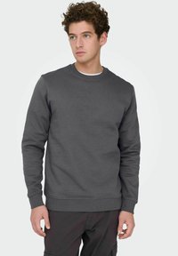 Homme aux cheveux bouclés portant un sweat-shirt à col rond gris foncé et un pantalon noir, regardant légèrement vers la gauche sur un fond clair uni.
