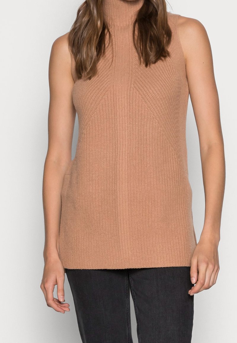 Maglione senza maniche color cammello con collo alto indossato da una persona con pantaloni scuri, in piedi contro uno sfondo bianco semplice.