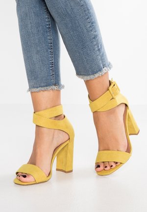 Žluté semišové sandály na vysokém podpatku s pásky kolem kotníku a otevřenou špičkou, s mohutným podpatkem a hladkou strukturou. Nositelná s krátkými džíny.