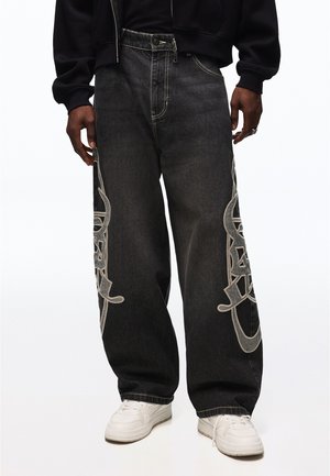 Personne portant un jean noir large avec de grands motifs abstraits gris clair sur les côtés, des baskets blanches et une veste noire.