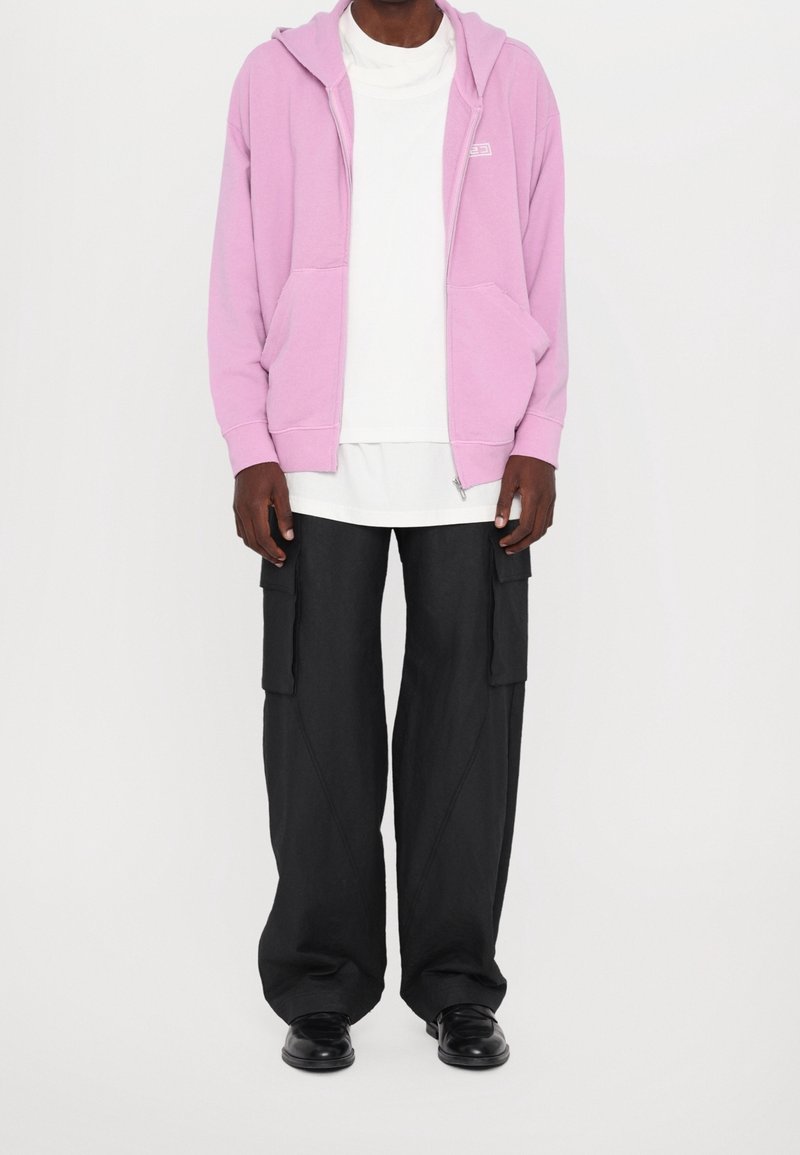 Homme portant un sweat à capuche zippé violet clair, un t-shirt blanc superposé, un pantalon cargo noir ample et des chaussures noires sur un fond blanc uni.