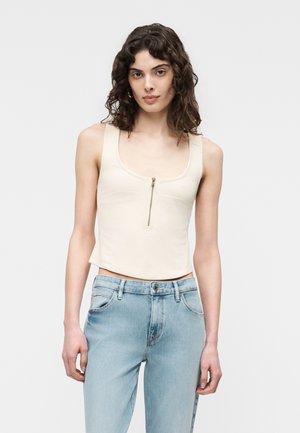 Młoda kobieta o kręconych włosach, ubrana w beżowy, bezrękawnik z zamkiem z przodu oraz jasnoniebieskie dżinsy z wysokim stanem, stoi na białym tle.