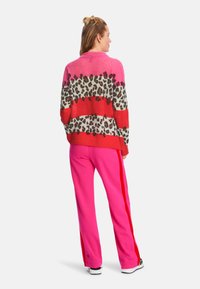 Bunt gestreifter Pullover in Pink-, Rot- und Tiermotiv-Design, kombiniert mit leuchtend rosa Hosen mit seitlichen Streifen, getragen mit weißen Sneakers.