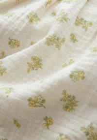 Blommig tyg med vit bakgrund, som har små gröna och gula blommönster. Materialet verkar mjukt och något texturerat.