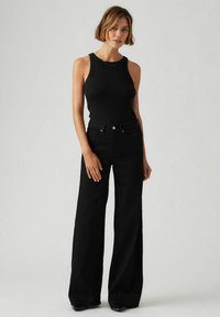 Débardeur noir à côtes avec une silhouette ajustée associé à un jean noir à taille haute et jambes larges. Détail de bouton en métal simple à la taille.