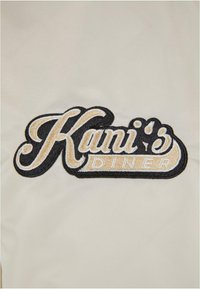 Gesticktes Patch mit dem Schriftzug "Kani's Diner" in geschwungener schwarzer und beiger Schrift auf einem cremefarbenen Hintergrund, der klar definierte Konturen und Texturen zeigt.