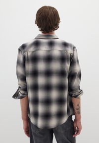 Camisa de franela a cuadros en blanco y negro con mangas enrolladas, que presenta una textura suave y un dobladillo redondeado. Tatuaje parcialmente visible en el brazo.