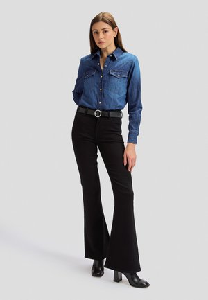 Žena koja nosi plavu jeans košulju s gumbima, crne trapez hlače s remenom i crne čizme s potpeticama, stoji na jednobojnoj svijetloj pozadini.