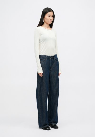 Witte blouse met lange mouwen en een ronde hals, gecombineerd met losvallende donkerblauwe denim jeans. Model draagt zwarte schoenen en minimale sieraden.