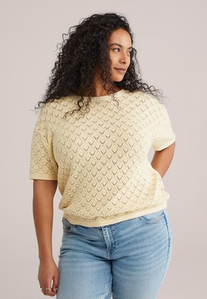 Kvinde med krøllet hår iført en cremefarvet kortærmet striktrøje og lyseblå jeans, der ser til siden med den ene hånd på hoften.