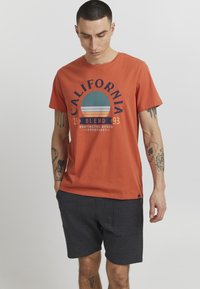 Narančasta pamučna majica s kružnim plavim i bijelim grafičkim uzorkom koji glasi "California Blend 1993" i tamnosive kratke hlače. Jednostavan dizajn stražnje strane.
