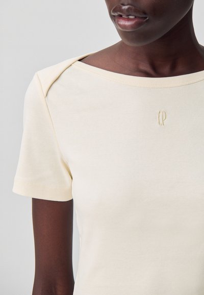 T-shirt beige chiaro con maniche corte, un ampio scollo e un logo ricamato sottile sul petto. Tessuto di cotone liscio con una forma aderente.