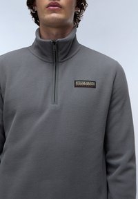 Sudadera de forro polar gris con diseño de media cremallera, que presenta un cuello alto y un pequeño parche con logo texturizado en el pecho. Textura suave y acogedora.