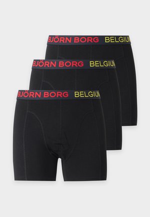 Homme athlétique debout pieds nus, portant un boxer noir avec une ceinture à logo, regardant vers sa gauche sur un fond blanc uni.