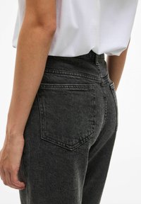 Crne traperice s visokim strukom, dizajnom s pet džepova i suptilnim izbljeđivanjem. Zatvoren prikaz leđa, koji prikazuje detalje šavova i teksturu.