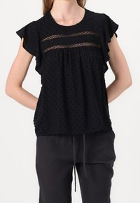 Blouse à pois noirs avec des manches courtes volantées et un détail en mesh transparent sur le buste. Légère, coupe décontractée, associée à un pantalon texturé foncé.