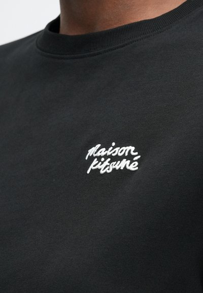 Černá mikina se bílým vyšitým logem "maison kitsuné" na levé hrudi, kterou nosí osoba viditelná od krku dolů.